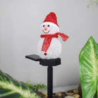 Solar snowman light with red hat and scarf, standing on a black pole among green leaves.